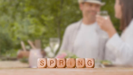 Concept-With-Wooden-Letter-Cubes-Or-Dice-Spelling-Spring-Against-Background-Of-Couple-Eating-Meal-Outdoors